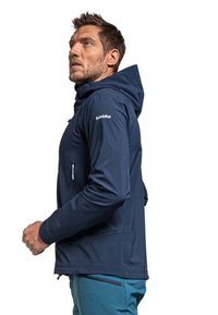 Marineblaue Softshell-Jacke mit Kapuze, mit Reißverschlusstaschen und Logodetail an der Schulter, kombiniert mit türkisfarbenen Hosen. Glatte Textur, taillierter Schnitt.