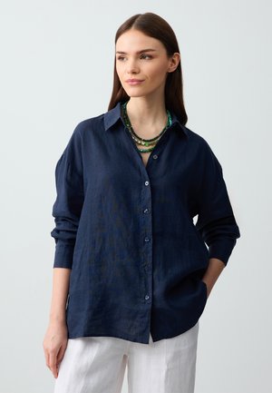 Camicia in lino blu navy con maniche lunghe, chiusura a bottoni e colletto classico. Abbinata a una collana di perline verdi a più strati.