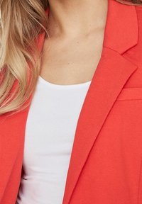 Rode blazer met een getailleerde pasvorm, gestructureerde stof en een enkele borstzak, gedragen over een wit tanktopje.