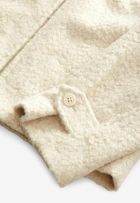 Tissu beige texturé avec une surface douce et duveteuse et un revers plié comportant un bouton beige. Fermeture éclair le long du centre.