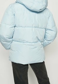 Lyseblå dunjakke med hætte, der har quiltede sektioner og en glat, skinnende overflade. Båret over mørke bukser, set bagfra.