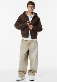 Bruine zip-up hoodie in een ontspannen pasvorm, gelaagd over een lichtblauw en wit gestreept shirt, in combinatie met lichtbeige baggy jeans en witte sneakers.
