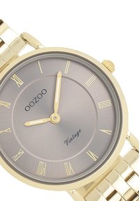 Montre-bracelet en ton doré avec un cadran rond gris, des chiffres romains et des aiguilles simples pour les heures et les minutes ; présente un bracelet en métal à maillons.