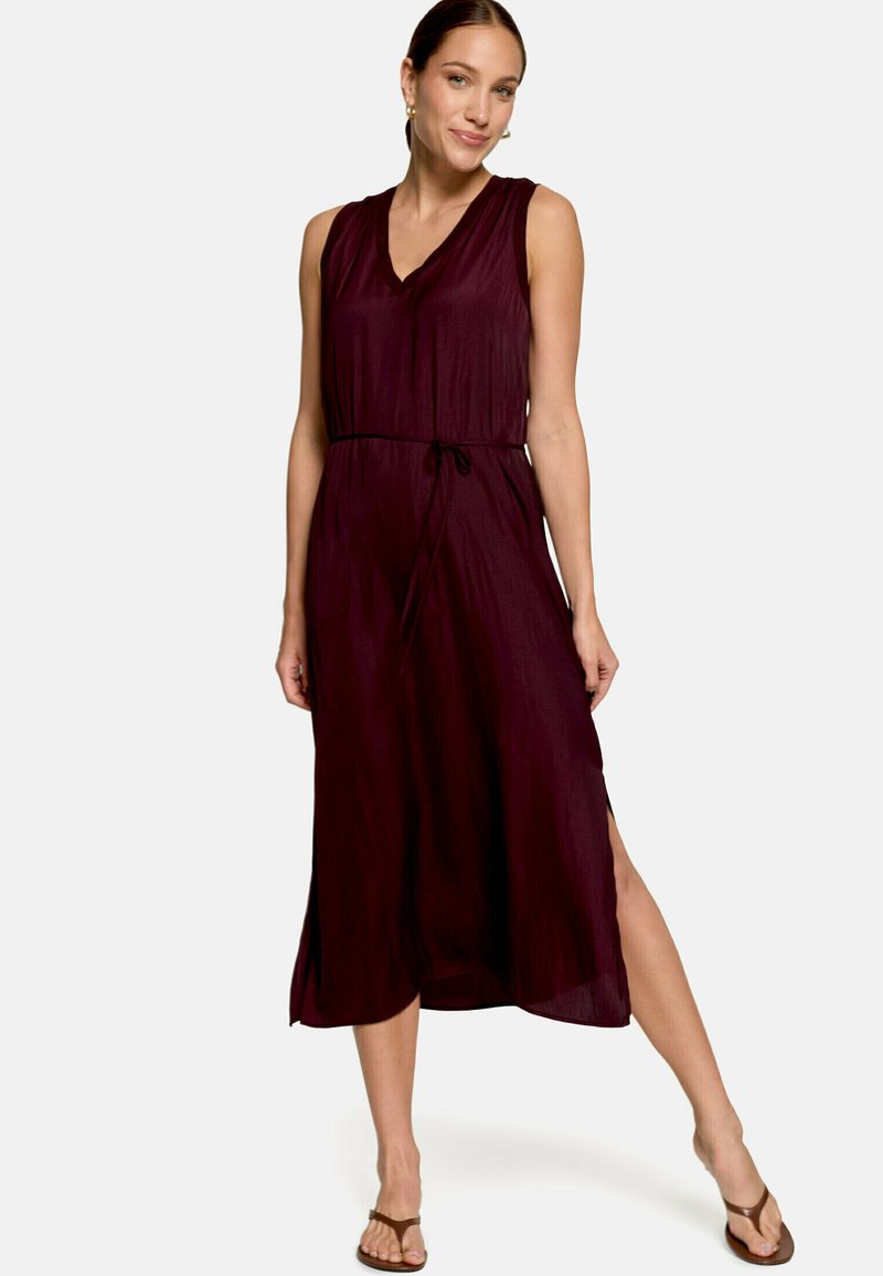 Femme portant une robe midi bordeaux foncé sans manches avec des fentes latérales, une ceinture à la taille et des sandales marron, debout avec une jambe tendue.