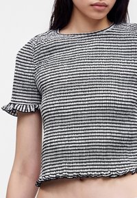 Γυναίκα φοράει μαύρο-άσπρο ριγέ, υφασμένο crop top με κοντά μανίκια και βολάν στα άκρα των μανικιών και του τελειώματος.