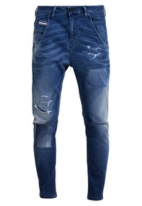 Modré džíny z denimu s roztřepenými detaily, obsahující více kapes, zip s lemem a zúžený střih. Osvětlené oblasti zvyšují texturu.