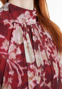 Blumenbluse mit hohem Kragen, ausgestattet mit Falten und einer Farbpalette aus tiefem Rot, Blush und Creme. Leichtes, leicht texturiertes Material.