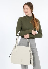 Jeune femme avec de longs cheveux en queue de cheval, portant un col roulé côtelé olive et un pantalon gris, tenant un grand sac à main blanc cassé avec des doubles poignées.