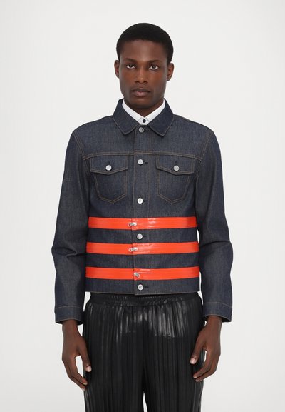 Veste en denim de couleur bleu foncé, avec des bretelles en plastique orange, des boutons en argent et deux poches poitrine ; associée à un pantalon noir plissé.