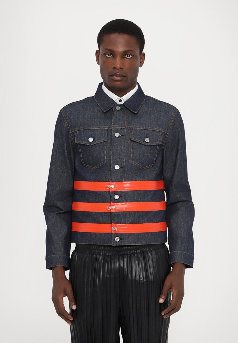 Veste en denim de couleur bleu foncé, avec des bretelles en plastique orange, des boutons en argent et deux poches poitrine ; associée à un pantalon noir plissé.