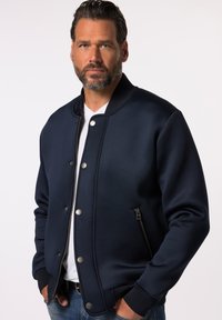 Marinfärgad bomberjacka med en mjuk textur, tryckknappar, ribbade ärmslut och en dragkedjeförsedd sidficka. Stylerad över en vit t-shirt.