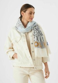 Lichtblauwe en grijs gemarmerde sjaal met franjes, gedragen rond de nek, boven een beige top en een witte denimjack.