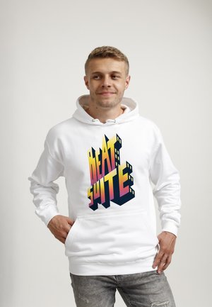 Młody mężczyzna w białej bluzie z kapturem z kolorową grafiką "BEAT SUITE", stoi z rękami w kieszeniach, patrząc lekko na bok.