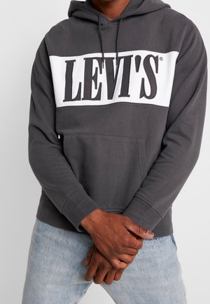 Mężczyzna ubrany w ciemnoszarą bluzę z kapturem Levi's z białym panelem i dużym logo, z rękami złożonymi przed sobą, noszący jasnoniebieskie dżinsy.