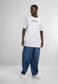 White cotton t-shirt with short sleeves featuring graphic detail. Paired with loose, dark blue denim pants and white sneakers.