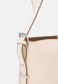 Sac à main en cuir crème avec une finition texturée, doté d'une bandoulière plate, d'accents métalliques argentés et d'un design simple et sans fermeture.