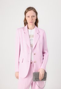 Blazer rosa claro con dos bolsillos frontales y botones dorados, combinado con pantalones a juego. La modelo sostiene un bolso metálico texturizado con una cadena.