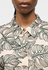 Εγγύς πλάνο ατόμου που φορά μπεζ πουκάμισο με πράσινα κεντητά σχέδια φύλλων και λαμπερά στρογγυλά κουμπιά, εμφανίζοντας μόνο το κάτω μέρος του προσώπου και τον λαιμό.