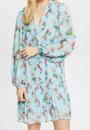 Femme portant une robe à manches longues bleu clair avec un motif floral rose et vert, jupe plissée, manches légèrement bouffantes et décolleté en V.