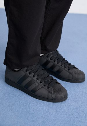 Czarne sneakersy Adidas Superstar z teksturowanym noskiem noszone z czarnymi spodniami na jasno-niebieskim dywanie.