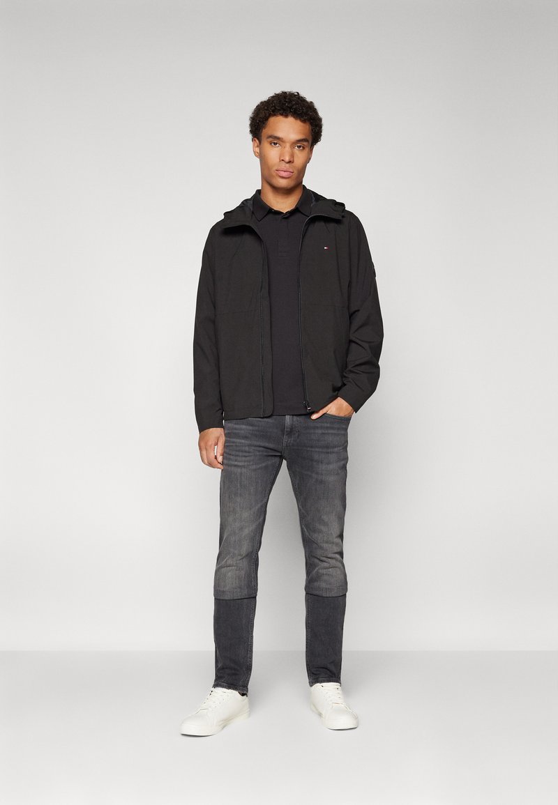 Chaqueta ligera negra con capucha, cierre de cremallera y dos bolsillos frontales, llevada sobre un polo negro, combinada con jeans grises y zapatillas blancas.