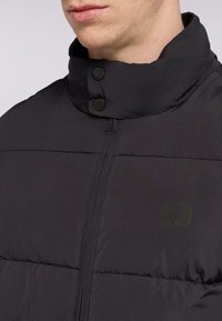 Svart puffjacka med hög krage, dragkedjestängning och två svarta tryckknappar. Slät textur med en rektangulär logotyp på bröstet.