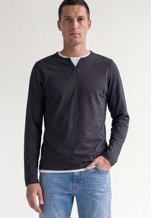 Uomo che indossa una camicia scura a maniche lunghe sopra una maglietta bianca e jeans azzurri, in piedi con una mano in tasca su sfondo uniforme.