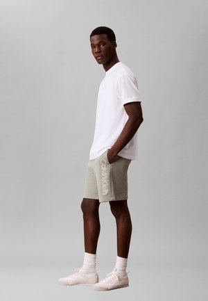 Hombre de pie con las manos en los bolsillos, vestido con camiseta blanca, pantalones cortos Calvin Klein gris claro, calcetines blancos y zapatillas blancas sobre fondo gris.