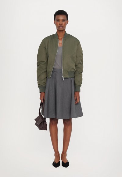 Olive green bomber jacket, gray top, gray pleated skirt, and black pointed-toe flats; holding a small brown bag. Simple design, textured fabric.