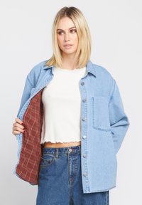 Denim jack met zilveren knopen, borstzak en geblokte bruine voering, gedragen over een witte crop top. Heeft een relaxte pasvorm.