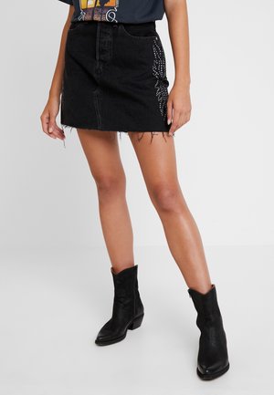 Person wearing a black denim mini skirt with studded side details and black mid-calf boots, standing against a plain background.