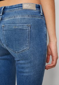 Jeans de mezclilla azul con un diseño ajustado, que presentan un bolsillo trasero con costura, una etiqueta de marca en la parte posterior y una textura suave.