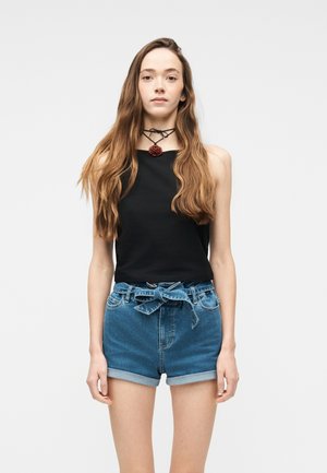 Νεαρή γυναίκα με μακριά κυματιστά μαλλιά, φοράει μαύρο αμάνικο τοπ, τζιν σορτς με δεμένη ζώνη και κολιέ τσόκερ με τριαντάφυλλο.