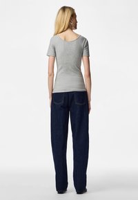 Kvinde med langt blondt hår iført en grå kortærmet top og mørkeblå vidde bukser, stående med ryggen til mod en hvid baggrund.