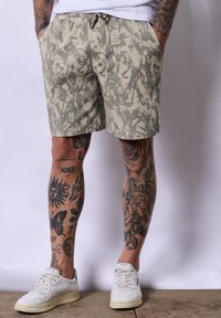 Beżowe szorty w wzory z teksturowanej powierzchni, z bocznymi kieszeniami i elastycznym pasem ze sznurkiem. Noszone z białymi trampkami.