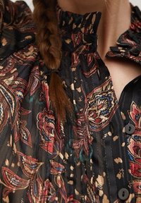 Blouse noire avec un motif floral paisley en rouge, bleu et crème. Présente une texture plissée et un col froncé avec des boutons.