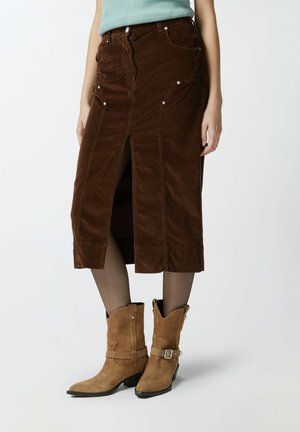 Jupe en velours côtelé marron avec une fente devant, des poches avant et des éléments en métal argenté. Associée à des bottes de cowboy en daim beige.