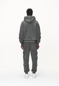 Man som står vänd bort med en mörkgrå hoodie och cargosbyxor med flera fickor samt vita sneakers mot en vit bakgrund.