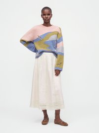 Femme portant un pull multicolore pastel, une longue jupe blanche avec bordure en dentelle et des ballerines marron à lacets, debout devant un fond blanc uni.
