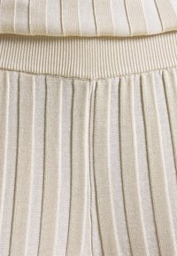 Béžová pletená tkanina s vertikálními skladbami, s žebrovanými akcenty a texturovaným povrchem, vytvářející vrstvený vzhled.