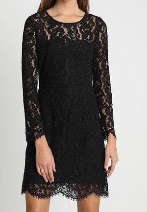 Vrouw draagt een nauwsluitende zwarte kanten jurk met lange doorschijnende mouwen en een geschulpte zoom, staand tegen een effen lichte achtergrond.