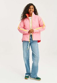 Růžová prošívaná bunda s žlutými akcenty a vysokým límcem, kombinovaná se světlými modrými džíny a zelenými teniskami s bílými tkaničkami.