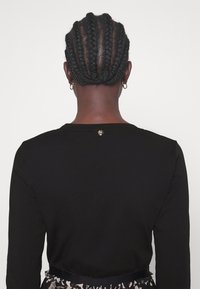 Personne vue de dos avec des cheveux tressés, portant de petites boucles d'oreilles anneaux et un haut noir à manches longues, avec une jupe à motifs visible à la taille.