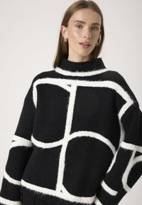 Suéter de lana negra con un cuello alto, que presenta patrones geométricos grandes en blanco y un acabado suave y texturizado. Mangas sueltas y de corte relajado.