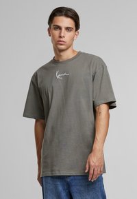 T-shirt en coton vert olive avec une coupe décontractée, des manches courtes et un logo manuscrit blanc sur la poitrine, associé à un jean en denim bleu.