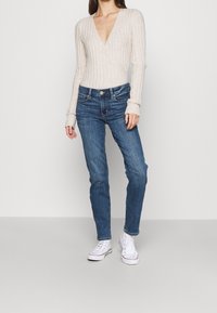 Světle béžový žebrovaný svetr s hlubokým V-výstřihem, kombinovaný s tmavě modrými slim-fit džíny a bílými plátěnými teniskami. Jednoduchý a čistý design.