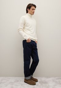 Cremefarbener Rollkragenpullover, dunkelblaue Jeans, braune Wildlederstiefel. Einfaches Design mit hochgekrempelten Ärmeln und sichtbaren Nahtdetails.