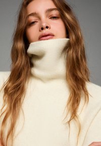 Krämfärgad polokrage-tröja i ribbat mönster, med hög krage och mjukt tyg. Håret faller ner i lätta vågor.