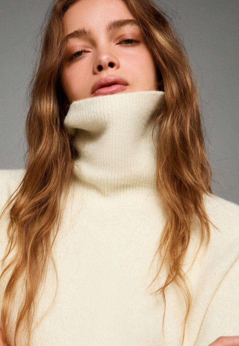 Krämfärgad polokrage-tröja i ribbat mönster, med hög krage och mjukt tyg. Håret faller ner i lätta vågor.