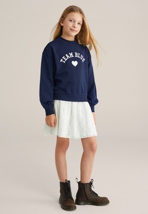 Granatowa bluza z napisem „TEAM BLUE” i grafiką serca, zestawiona z białą koronkową spódnicą i brązowymi sznurowanymi botkami z czarnymi akcentami.
